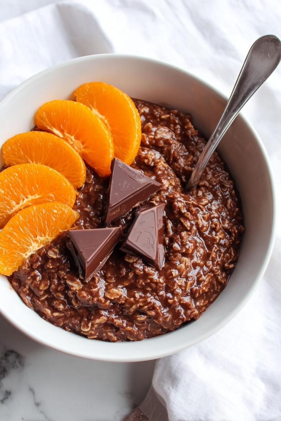 The image shows a white bowl filled with a thick, dark brown chocolate oatmeal mixture that has a slightly rough texture from the oats. On the top left side of the oatmeal, there are six orange slices arranged in a neat cluster, while on the top right side, two triangular pieces of rich, dark chocolate rest partially melted on the oatmeal's surface. A silver spoon is placed inside the bowl on the right side, angled upward. The bowl sits on a white marbled surface with part of a white cloth visible in the background. Photo taken with an iphone --ar 2:3 --v 7 - Chocolate Orange Porridge, breakfast with chocolate and orange, quick healthy breakfast recipes, cozy morning porridge, citrus and cocoa oatmeal