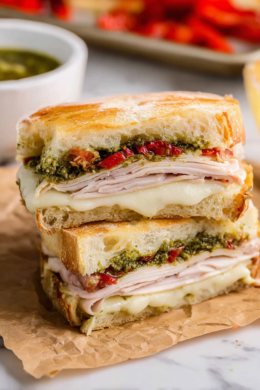 The image shows a sandwich cut in half and stacked, placed on crinkled brown paper against a white marbled surface. The sandwich has three main layers of fillings. The bottom layer consists of melted white cheese with a soft texture. Above it is a layer of thin, light pink slices of turkey. On top of the turkey, there is a green layer of pesto sauce mixed with small pieces of red roasted peppers. The sandwich is held together by two thick slices of golden brown, slightly crusty bread with a soft inside, forming the top and bottom of the sandwich. In the background, there is a white bowl with green sauce and part of a baking tray with more red roasted peppers visible. Photo taken with an iphone --ar 2:3 --v 7 - Copycat Starbucks Turkey Pesto Sandwich, Starbucks turkey pesto sandwich copycat, homemade Starbucks turkey pesto, how to make turkey pesto sandwich, best turkey pesto sandwich