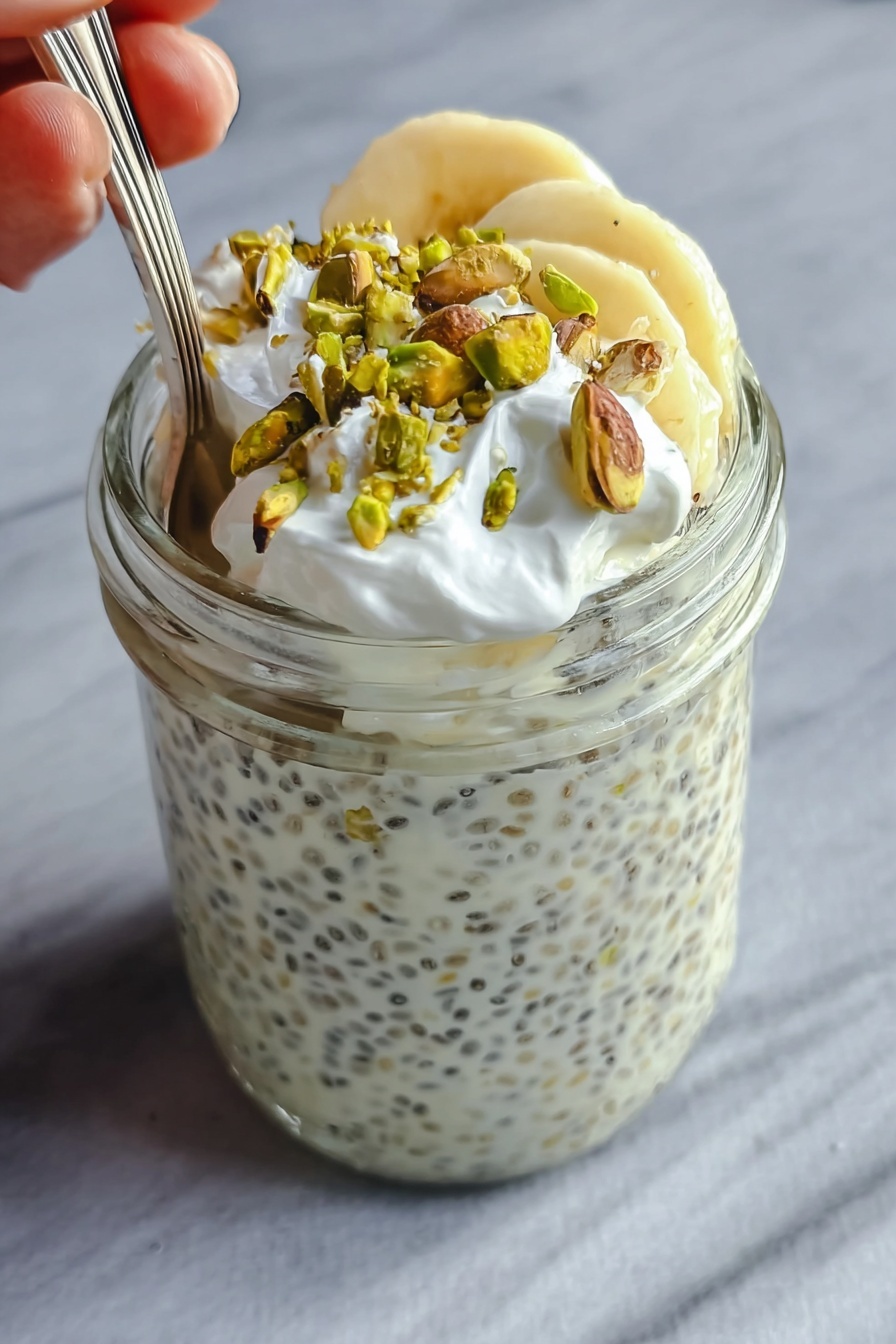 The image shows a clear glass jar filled almost to the top with a creamy white chia pudding that has small black chia seeds spread evenly throughout. On the top layer inside the jar, there are a few slices of light yellow banana, which are partially covered by the creamy pudding. Over the bananas, there is a layer of small white whipped cream dollops, adding a soft texture. Scattered on the very top are roughly chopped green pistachio nuts that add a contrasting color and crunchy texture. A spoon is placed inside the jar, with a woman's hand holding it gently from the left side. The jar is set against a smooth, white marbled surface. Photo taken with an iphone --ar 2:3 --v 7 - Creamy Pistachio Protein Overnight Oats, healthy overnight oats, high-protein breakfast, easy breakfast ideas, pistachio yogurt oats