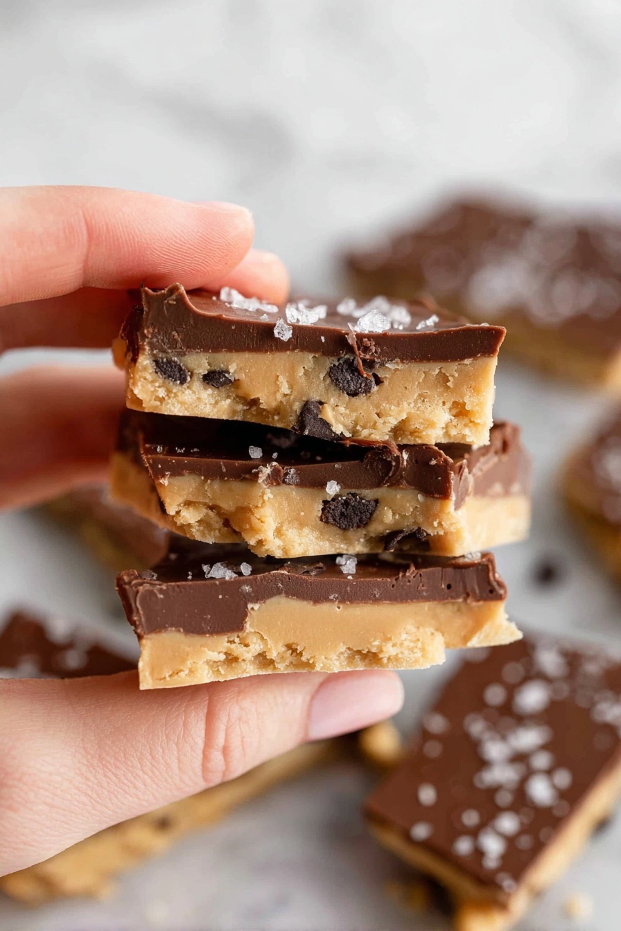 A woman's hand is holding three pieces of snack bark stacked closely together. Each piece has two layers: a bottom light tan layer with a soft texture and dark chocolate chip bits mixed in, and a thick top layer of smooth, dark brown chocolate. The top chocolate layer is sprinkled with small flakes of white sea salt. More broken pieces of the same snack are scattered on a white marbled surface in the background, showing both tan and dark chocolate layers clearly. Photo taken with an iphone --ar 2:3 --v 7 - Healthy Cookie Dough Bark, healthy cookie dough bark recipe, guilt-free cookie dough snack, easy healthy dessert, no-bake cookie bark