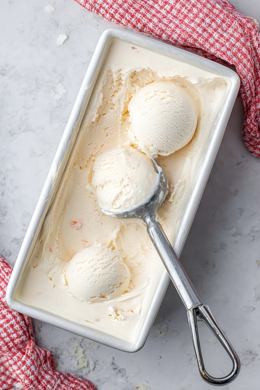 A white metal rectangular container filled with creamy, pale beige ice cream shows two round scoops placed near the top left corner. Ice cream has a smooth but slightly textured surface with small visible bits inside. A silver ice cream scooper rests partially buried on the right side, with some ice cream residue on it. A white and red checkered cloth is partly visible in the upper right corner on a white marbled surface. photo taken with an iphone --ar 2:3 --v 7 - Homemade Coconut Ice Cream, easy coconut ice cream without ice cream maker, tropical coconut ice cream recipe, dairy-free coconut ice cream, quick homemade coconut ice cream