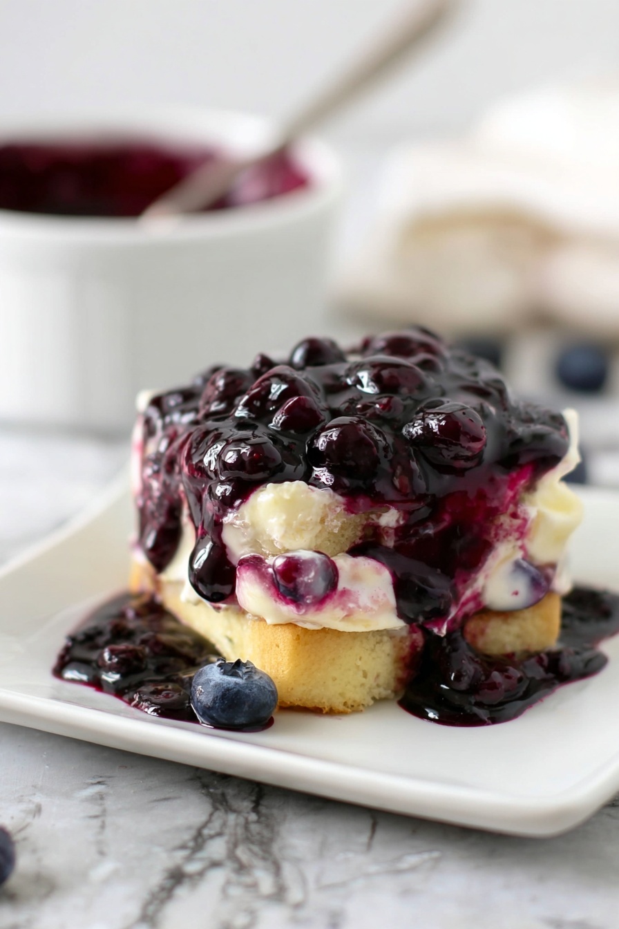 The image shows a small dessert on a white square plate on a white marbled surface. The dessert has two main layers of light golden cake, between which there is a creamy white filling mixed with some purple blueberry syrup. On top, a thick layer of dark purple blueberry sauce with whole blueberries covers the cake, dripping down the sides and pooling on the plate. In the background, there is a blurred white bowl with more dark purple blueberry sauce and a spoon. The scene has a soft natural light. photo taken with an iphone --ar 2:3 --v 7 - Blueberry Syrup for Waffles and Pancakes, blueberry syrup recipe, homemade blueberry syrup, fruit topping for breakfast, easy blueberry syrup