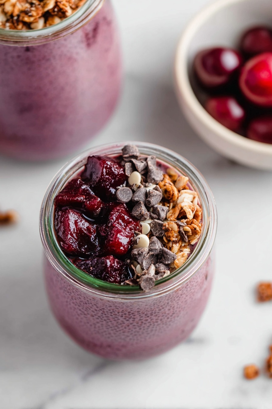 The image shows a small glass jar filled with three distinct layers from bottom to top: a smooth, thick purple chia pudding base with a slightly bumpy texture, topped on one side with dark red cherry chunks that look juicy and glossy, and on the other side with crunchy light brown granola mixed with dark brown chocolate chips, all neatly arranged. Above the jar, part of another jar is visible filled with purple chia pudding, and to the right, a white bowl with whole dark red cherries on a white marbled surface. Photo taken with an iphone --ar 2:3 --v 7 - Cherry Chia Pudding, healthy chia seed pudding with cherries, easy cherry chia pudding, nutritious fruit chia pudding, quick breakfast cherry pudding