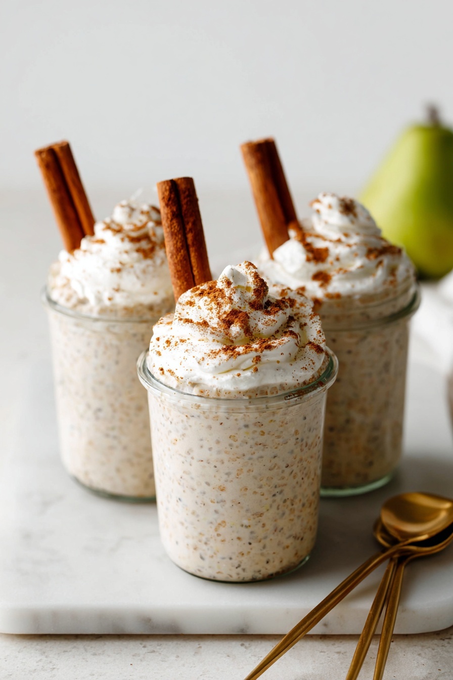 Three small clear glass jars are filled with a creamy, speckled beige mixture, each topped with a dollop of white whipped cream sprinkled with brown cinnamon powder. Two brown cinnamon sticks are inserted upright into the whipped cream on each jar, leaning slightly against each other. The jars are placed close together on a white marbled surface. To the side, two shiny gold spoons rest on the same surface. In the blurry background, a green fruit is partially visible. photo taken with an iphone --ar 2:3 --v 7 - Chai Spiced Overnight Oats, healthy chai oats, cozy breakfast recipes, nutritious overnight oats, easy breakfast ideas