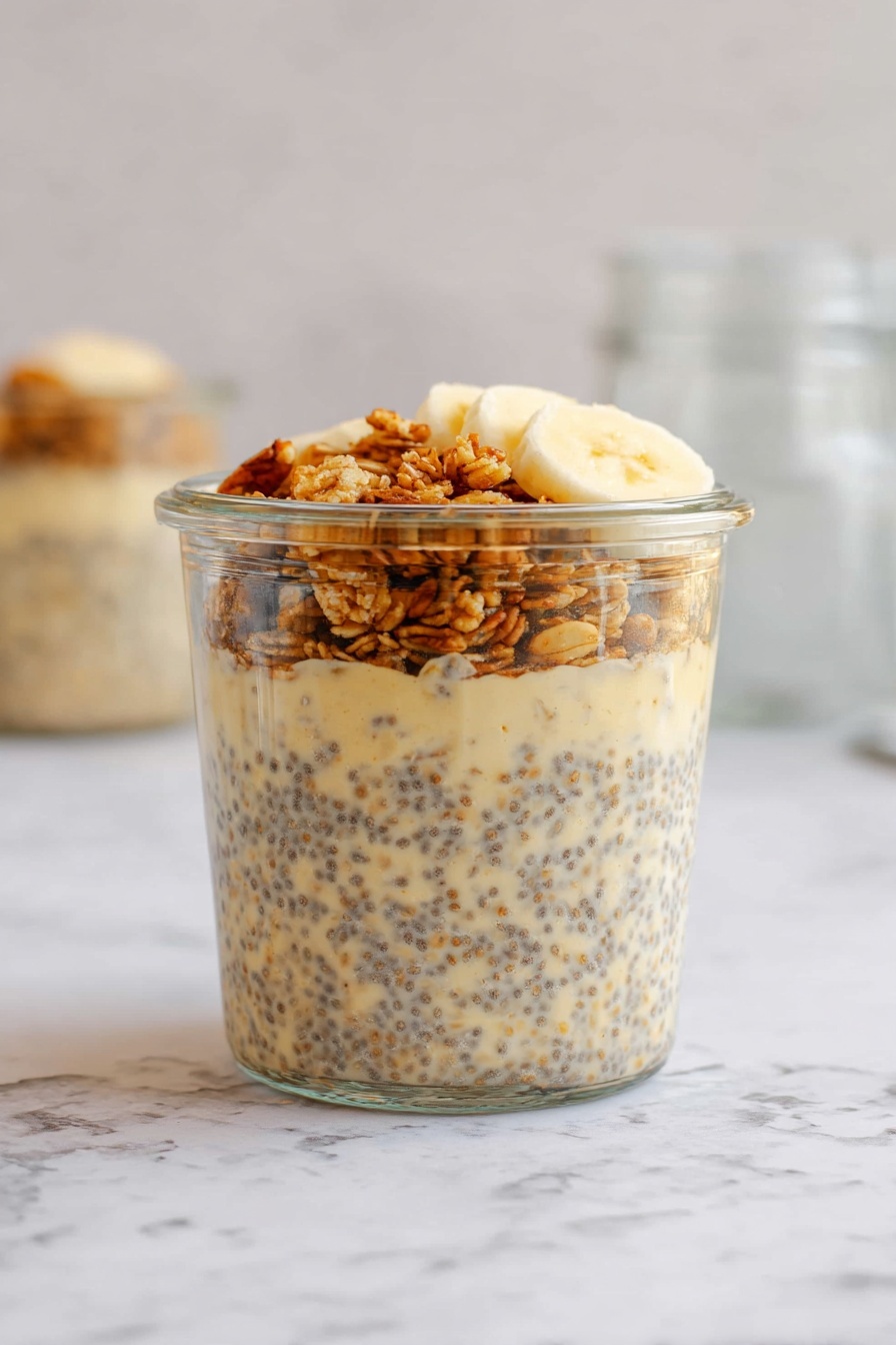 The image shows a clear glass jar filled with a three-layered breakfast dish. The bottom two layers consist of a creamy mixture with visible chia seeds and oatmeal that is light beige with dark specks. The top layer is a small portion of granola, golden brown in color, and a few slices of banana, pale yellow. The jar sits on a white marbled surface, with a blurred, light background and glass jars in the back. Photo taken with an iphone --ar 2:3 --v 7 - Brown Sugar Overnight Oats, healthy overnight oats, easy breakfast recipes, sweet breakfast ideas, protein-packed overnight oats